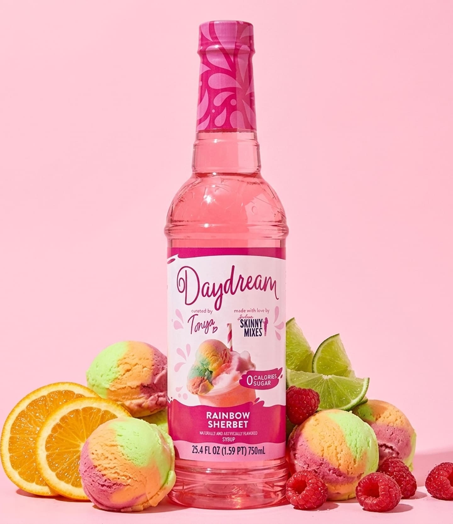 A bottle of Daydream Tropicals Rainbow Sherbet non-alcoholic mixer syrup, styled with colorful ice cream scoops, raspberries, limes, and orange slices on a solid pink background.